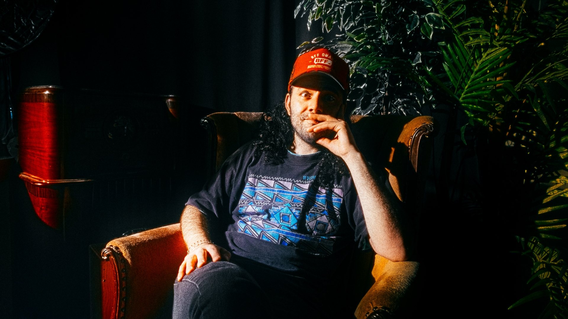 Kenny sitting in a velvet armchair under a spotlight, wearing a red cap